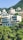 A modern, multi-story building nestled amongst lush green trees and set against a mountainous backdrop. The building features large windows and balconies, with a minimalist architectural design. Surrounding the main building are other residential structures set on the hillside.