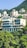 A modern, multi-story building nestled amongst lush green trees and set against a mountainous backdrop. The building features large windows and balconies, with a minimalist architectural design. Surrounding the main building are other residential structures set on the hillside.