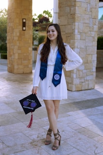 A young woman standing in a well-lit outdoor setting, wearing a white dress and a blue graduation stole. She is holding a graduation cap decorated with the words 'It's all happening' and a pair of sunglasses. The background consists of stone pillars and green foliage.