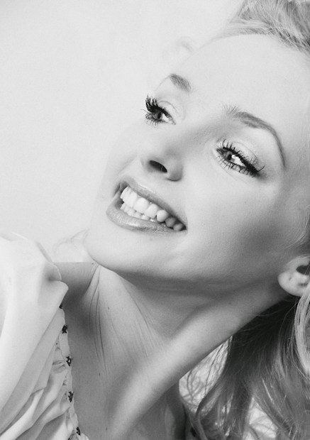 A joyful black and white close-up portrait of a smiling woman with bright eyes and long eyelashes, gazing upward. Her hair appears to be styled neatly, and she is wearing a light-colored garment.