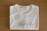 A folded white T-shirt with a stylish logo printed in black on the front. The brand label at the neckline reads 'Vanny Clothing Co.' The shirt lies on a wooden surface, which serves as a simple backdrop.