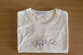 A folded white T-shirt with a stylish logo printed in black on the front. The brand label at the neckline reads 'Vanny Clothing Co.' The shirt lies on a wooden surface, which serves as a simple backdrop.