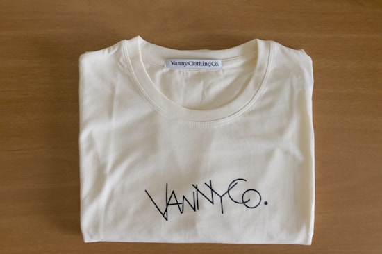 A folded white T-shirt with a stylish logo printed in black on the front. The brand label at the neckline reads 'Vanny Clothing Co.' The shirt lies on a wooden surface, which serves as a simple backdrop.