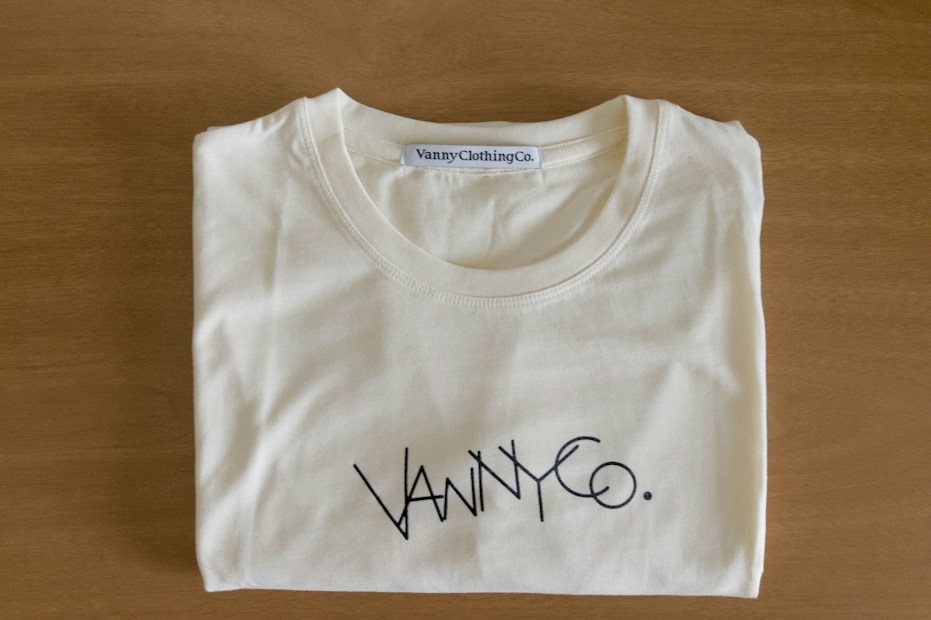 A folded white T-shirt with a stylish logo printed in black on the front. The brand label at the neckline reads 'Vanny Clothing Co.' The shirt lies on a wooden surface, which serves as a simple backdrop.