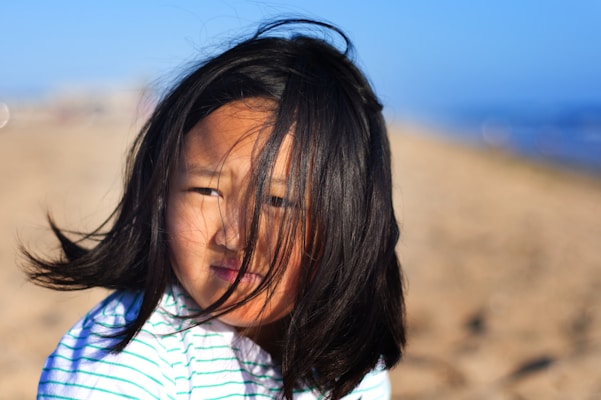 A young girl with long black hair looks pensively at the camera, as her hair is gently blown by the wind. The background features a sandy beach with the ocean visible in the distance, and the scene is bathed in warm sunlight.