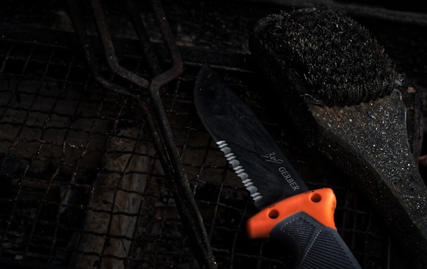 A serrated knife with an orange and black handle lies on a grill grate, alongside a used barbecue grill brush with bristles. The scene is dark, with a focus on the textures of the metal and bristles, creating a rustic and rugged feel.