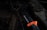 A serrated knife with an orange and black handle lies on a grill grate, alongside a used barbecue grill brush with bristles. The scene is dark, with a focus on the textures of the metal and bristles, creating a rustic and rugged feel.