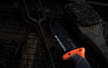 A serrated knife with an orange and black handle lies on a grill grate, alongside a used barbecue grill brush with bristles. The scene is dark, with a focus on the textures of the metal and bristles, creating a rustic and rugged feel.