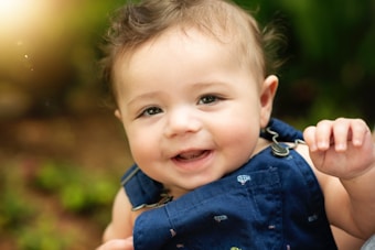 A smiling baby with curly brown hair wearing a blue outfit. The background is blurred, highlighting the baby's joyful expression and creating a warm atmosphere.
