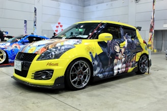 A customized yellow car displayed in an indoor exhibition setting, featuring vibrant anime-style artwork on its exterior. The design includes characters and colorful graphics with Japanese text. The setting includes other decorated cars and promotional materials in the background.