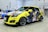 A customized yellow car displayed in an indoor exhibition setting, featuring vibrant anime-style artwork on its exterior. The design includes characters and colorful graphics with Japanese text. The setting includes other decorated cars and promotional materials in the background.