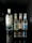 A lineup of four beverage bottles arranged in a row, with one large bottle on the left and three smaller bottles on the right. The large bottle is gin, while the smaller ones are Fever-Tree mixers, including ginger beer and tonic waters. The bottles are placed on a reflective black surface, creating a clear mirror reflection. The surroundings are dimly lit, which highlights the bottles and their reflections.
