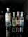 A lineup of four beverage bottles arranged in a row, with one large bottle on the left and three smaller bottles on the right. The large bottle is gin, while the smaller ones are Fever-Tree mixers, including ginger beer and tonic waters. The bottles are placed on a reflective black surface, creating a clear mirror reflection. The surroundings are dimly lit, which highlights the bottles and their reflections.