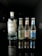 A lineup of four beverage bottles arranged in a row, with one large bottle on the left and three smaller bottles on the right. The large bottle is gin, while the smaller ones are Fever-Tree mixers, including ginger beer and tonic waters. The bottles are placed on a reflective black surface, creating a clear mirror reflection. The surroundings are dimly lit, which highlights the bottles and their reflections.