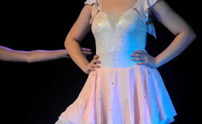 A person wearing a delicate ballet dress featuring a fitted bodice with decorative embellishments and a flowing skirt. The dress has a soft pink hue and is adorned with subtle sparkles. The background is dark, emphasizing the bright colors of the attire and the performer’s pose.