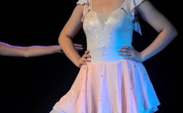 A person wearing a delicate ballet dress featuring a fitted bodice with decorative embellishments and a flowing skirt. The dress has a soft pink hue and is adorned with subtle sparkles. The background is dark, emphasizing the bright colors of the attire and the performer&rsquo;s pose.