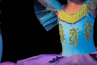 A close-up view of a person wearing a bright, colorful ballet costume with intricate details. The costume features a vibrant mix of blue and yellow, with shimmering embellishments that catch the light. The top part of the costume has ornamental decorations, while sheer fabric adds layers to the design.