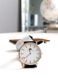 A stylish wristwatch with a minimalist design, featuring a white dial and gold markers. The watch has a black leather strap and is positioned standing on a white surface. In the background, a blurred globe and a brown object can be seen, adding to the aesthetic and sophisticated ambiance.