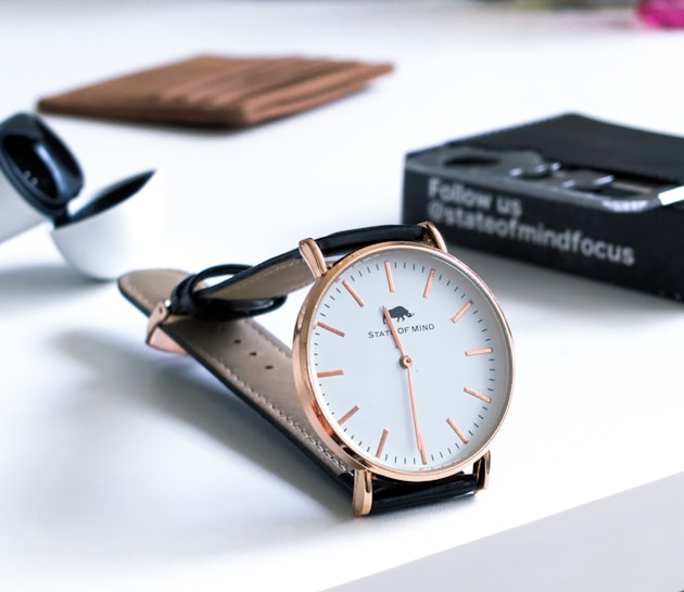 A stylish watch with a round dial and rose gold accents lies on a white surface. The watch has a simple, elegant design with minimalistic hour markers and a black leather strap. In the background, a pair of wireless earbuds and a black box with branding text can be seen.