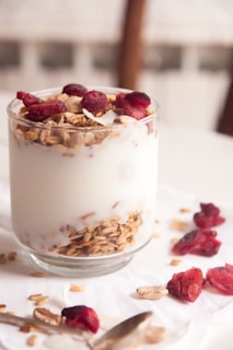 A glass filled with creamy yogurt, topped with layers of granola and pieces of dried red fruit, possibly cranberries or raspberries. The granola and fruit are also scattered on the white surface around the glass, alongside a spoon.