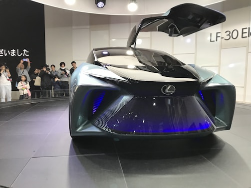 A futuristic concept car with a sleek, aerodynamic design is displayed. The car features a glossy black surface with blue lighting accents, and its gull-wing doors are open. Onlookers in the background are taking pictures, and the scene takes place at what appears to be an auto show, suggested by the signage and lighting.