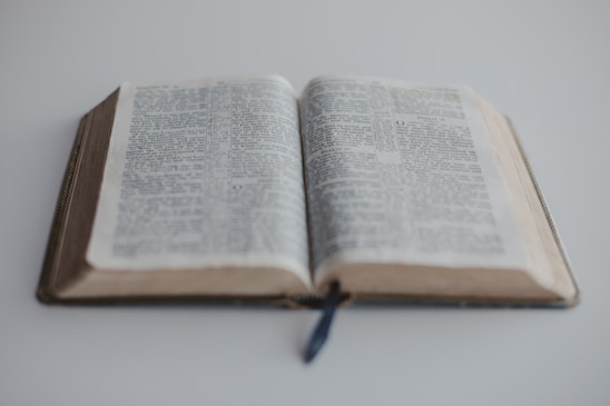 An open book resting on a white surface with a visible ribbon bookmark in the center. The focus is on the text on the right page, while the left page appears slightly out of focus. The book's pages have a slightly aged appearance.