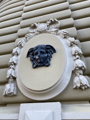A detailed sculptural decoration featuring a dark blue Medusa head set against a cream-colored oval background, surrounded by intricate floral and ribbon motifs. The surrounding structure is composed of horizontal ridges in a beige tone, emphasizing the ornate centerpiece.