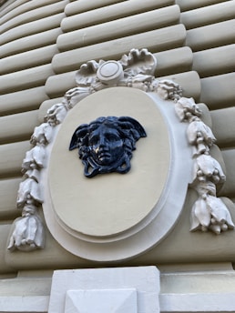 A detailed sculptural decoration featuring a dark blue Medusa head set against a cream-colored oval background, surrounded by intricate floral and ribbon motifs. The surrounding structure is composed of horizontal ridges in a beige tone, emphasizing the ornate centerpiece.
