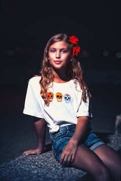 A young girl with long blonde hair wearing a white T-shirt featuring three colorful skulls, sitting on a stone bench at night. She has a red flower in her hair and is gazing directly at the camera.