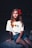 A young girl with long blonde hair wearing a white T-shirt featuring three colorful skulls, sitting on a stone bench at night. She has a red flower in her hair and is gazing directly at the camera.