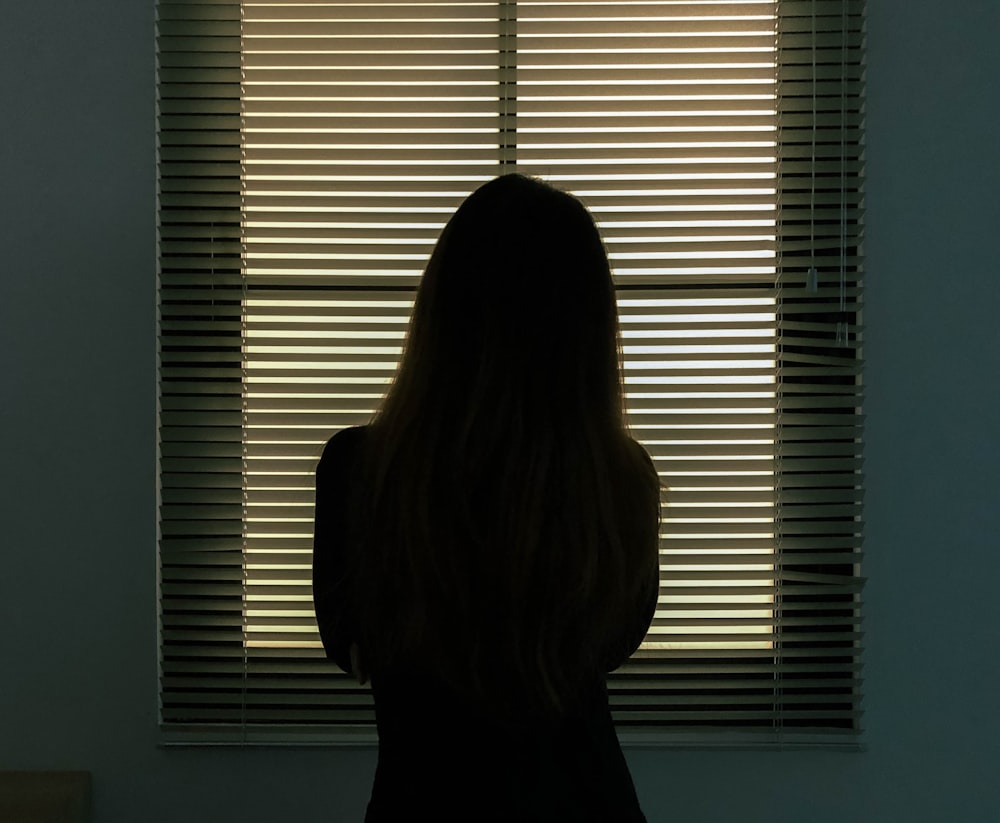 mulher na camisa preta de pé perto das persianas da janela