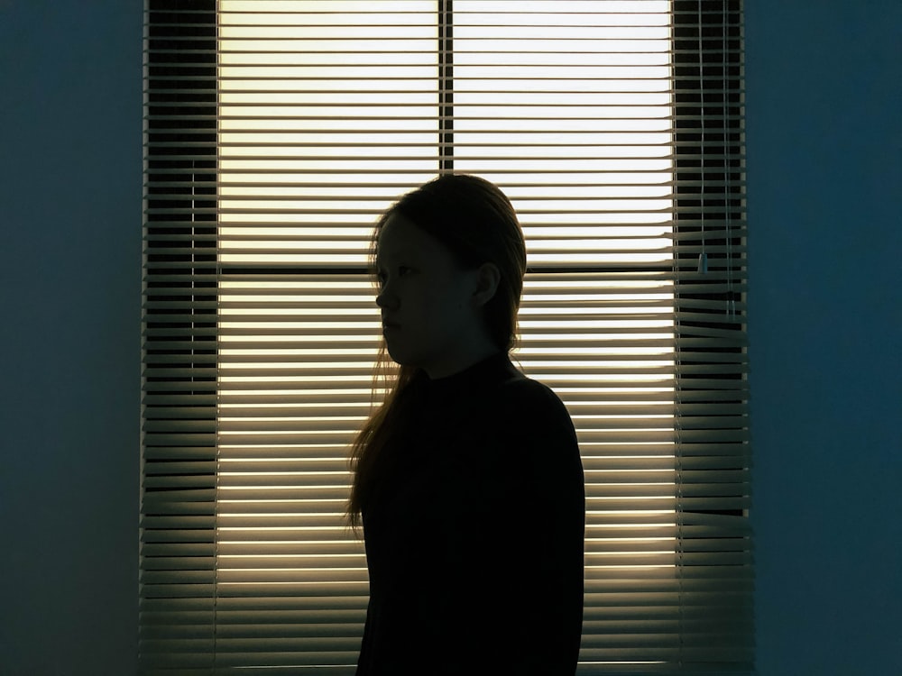 mulher na camisa preta de manga comprida de pé ao lado das persianas da janela