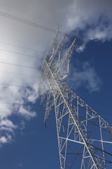 A tall metal power transmission tower stands against a backdrop of a partially cloudy blue sky. Power lines extend outwards from the tower, creating a network of cables in the sky.