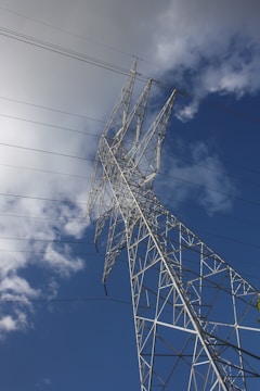 A tall metal power transmission tower stands against a backdrop of a partially cloudy blue sky. Power lines extend outwards from the tower, creating a network of cables in the sky.