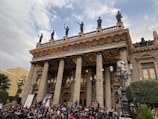 A grand neoclassical theater with tall columns and statues on the rooftop. A large crowd of people is gathered at the front steps, possibly for an event or performance. The building has ornate details and multiple street lamps are visible.