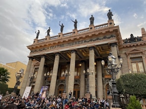 A grand neoclassical theater with tall columns and statues on the rooftop. A large crowd of people is gathered at the front steps, possibly for an event or performance. The building has ornate details and multiple street lamps are visible.