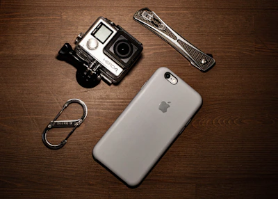 A collection of gadgets placed on a wooden surface including a GoPro camera, a smartphone with an Apple logo, a carabiner clip, and a multitool.