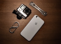A collection of gadgets placed on a wooden surface including a GoPro camera, a smartphone with an Apple logo, a carabiner clip, and a multitool.