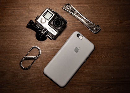 A collection of gadgets placed on a wooden surface including a GoPro camera, a smartphone with an Apple logo, a carabiner clip, and a multitool.