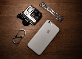 A collection of gadgets placed on a wooden surface including a GoPro camera, a smartphone with an Apple logo, a carabiner clip, and a multitool.