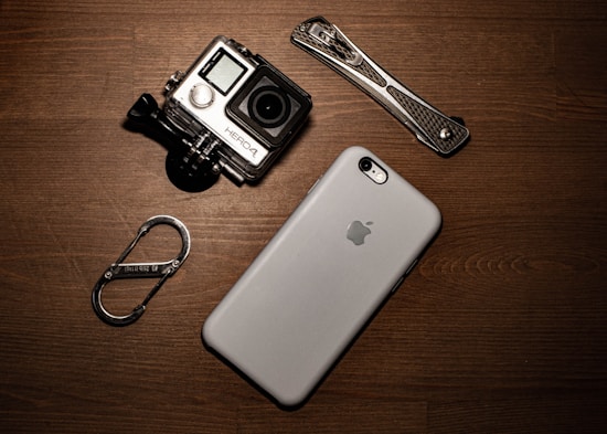 A collection of gadgets placed on a wooden surface including a GoPro camera, a smartphone with an Apple logo, a carabiner clip, and a multitool.