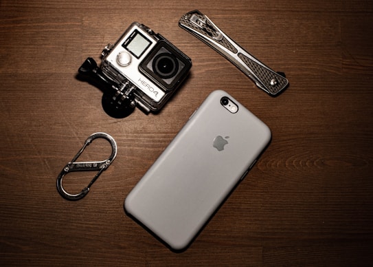 A collection of gadgets placed on a wooden surface including a GoPro camera, a smartphone with an Apple logo, a carabiner clip, and a multitool.