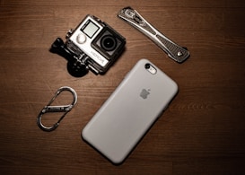 A collection of gadgets placed on a wooden surface including a GoPro camera, a smartphone with an Apple logo, a carabiner clip, and a multitool.