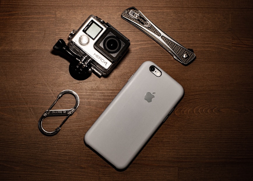 A collection of gadgets placed on a wooden surface including a GoPro camera, a smartphone with an Apple logo, a carabiner clip, and a multitool.