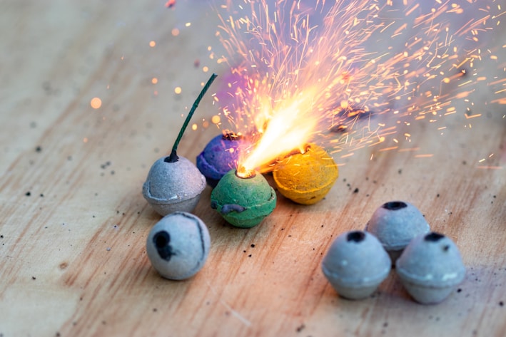 A collection of small spherical fireworks is arranged on a wooden surface. One of the fireworks is lit, emitting bright sparks and a burst of light. The spheres are in various colors, including gray, green, blue, and yellow, some with visible fuses.