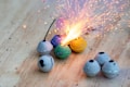 A collection of small spherical fireworks is arranged on a wooden surface. One of the fireworks is lit, emitting bright sparks and a burst of light. The spheres are in various colors, including gray, green, blue, and yellow, some with visible fuses.