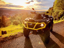 A rugged ATV with a bright yellow bumper is parked on a gravel path, facing a lush, sunlit valley. The landscape is filled with rolling green hills and scattered trees, with a vibrant sunset casting warm hues over the scene.