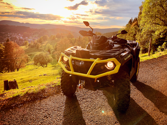 A rugged ATV with a bright yellow bumper is parked on a gravel path, facing a lush, sunlit valley. The landscape is filled with rolling green hills and scattered trees, with a vibrant sunset casting warm hues over the scene.