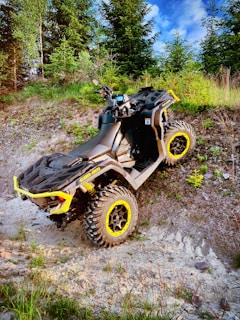 A rugged all-terrain vehicle with yellow accents is parked on a rocky, inclined surface surrounded by lush greenery and trees. The vehicle's large tires and sturdy design suggest it is equipped for off-road adventures.