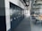 A spacious office or industrial storage space with large metal cabinets lined up along the wall, featuring circular locks. The area also includes modern office chairs and workstations with orange accents. Overhead, there is an array of exposed pipes and ventilation ducts.